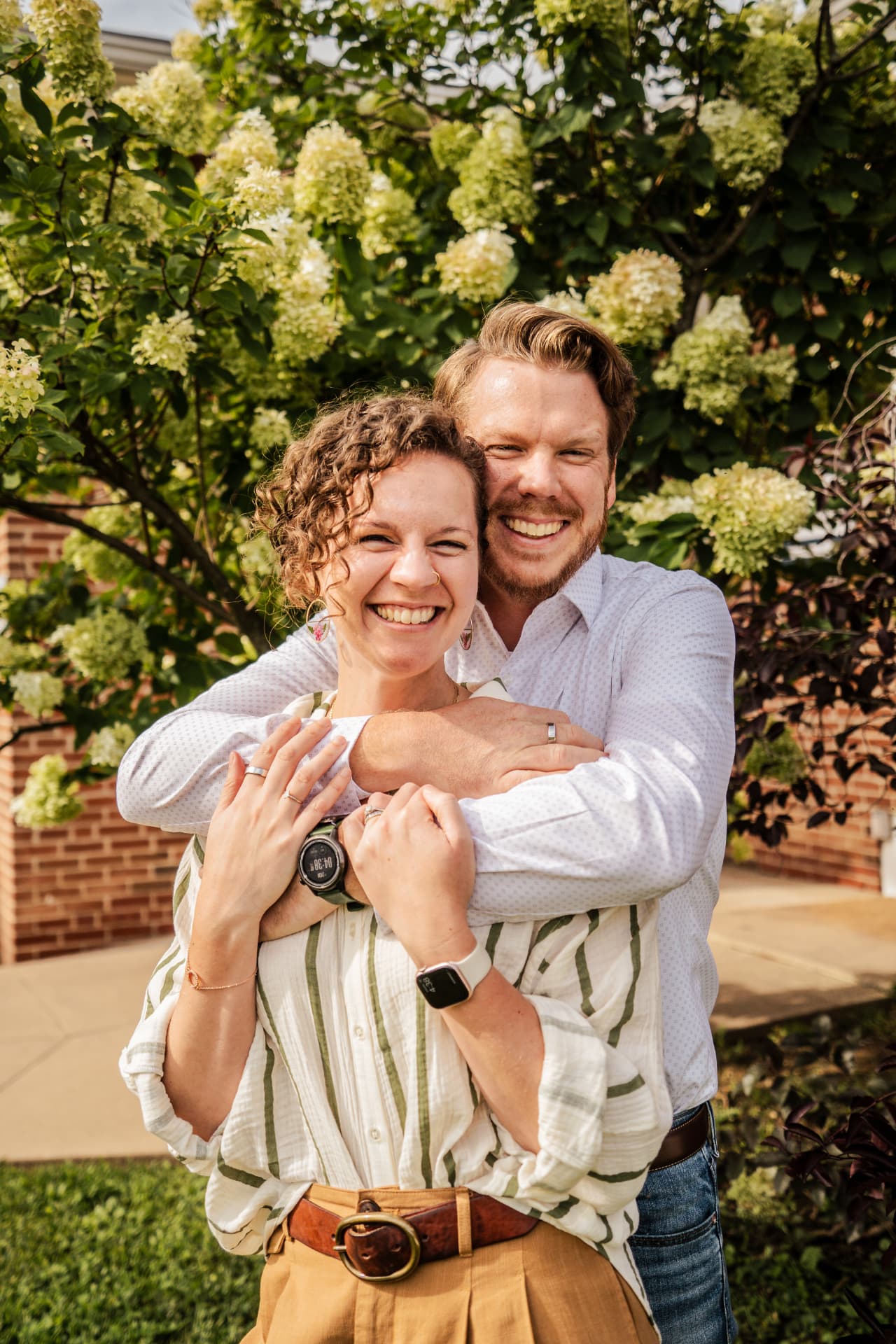 Brice and his wife smiling together outdoors in front of white hydrangea blooms